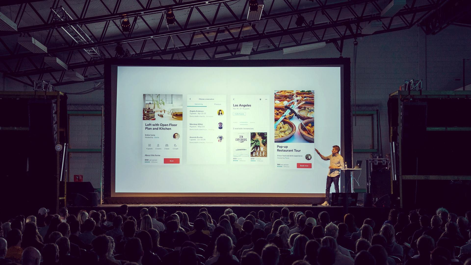 person discussing while standing in front of a large screen in front of people inside dim-lighted room